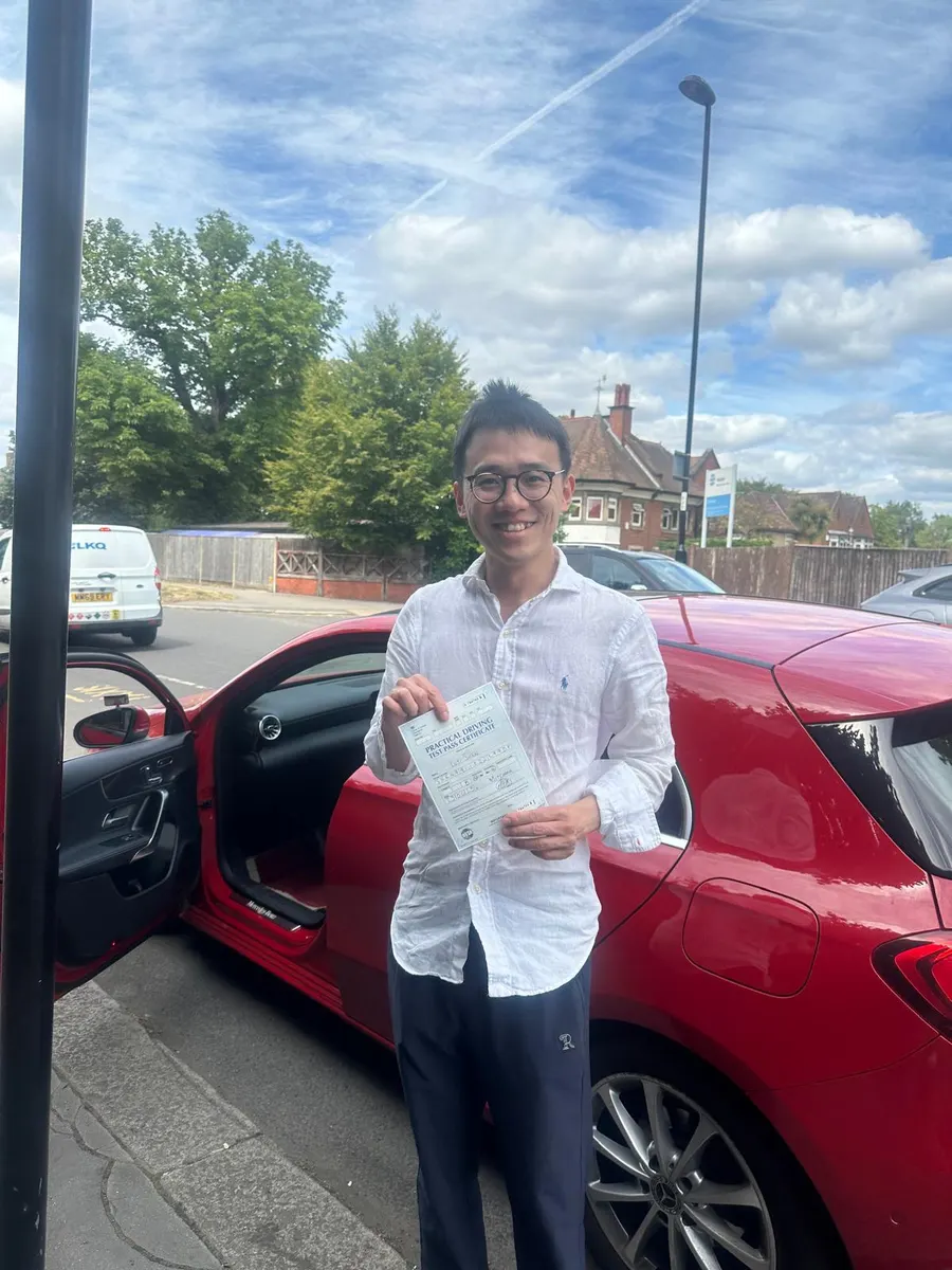 Young man in a white shirt holding his driving test pass certificate by the open door of a red Mercedes tuition car