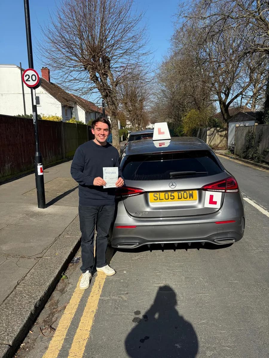 New driver holding his pass certificate beside a grey Mercedes tuition car on a tree-lined 20mph street